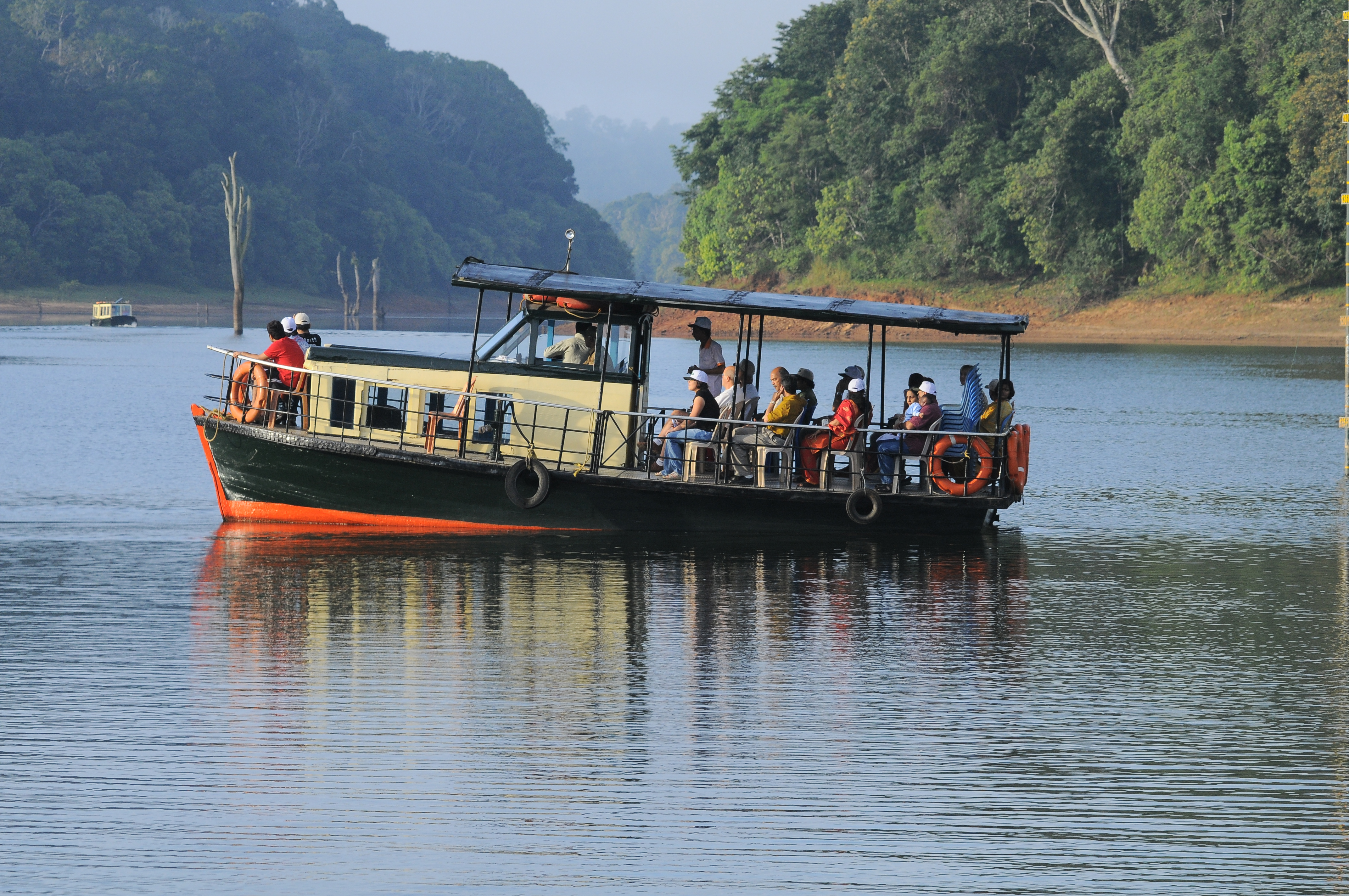 Tourists on a boat safari gliding through the serene Periyar Lake inside the Periyar Tiger Reserve, with forested hills reflected in the calm water