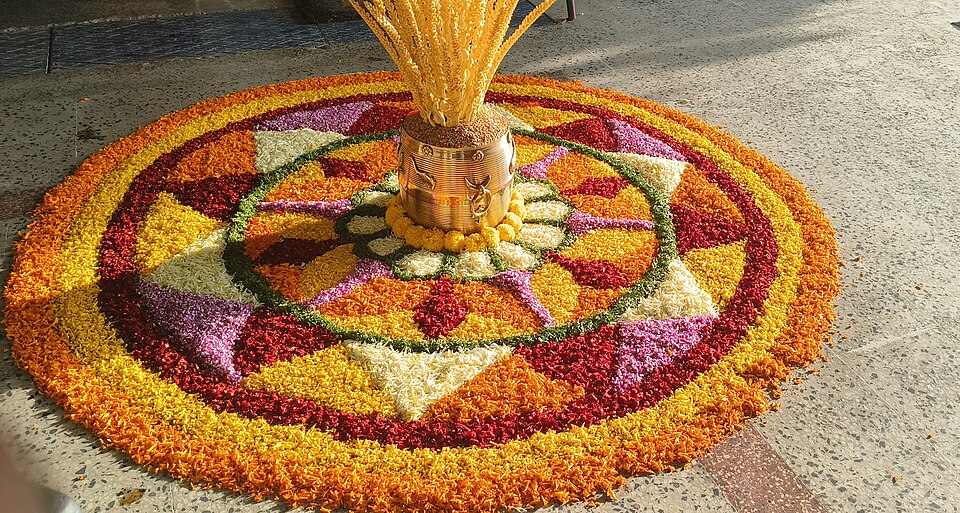 A vibrant, traditional flower carpet (Onappookkalam) arranged beautifully alongside a traditional brass Nirapara filled with paddy husk